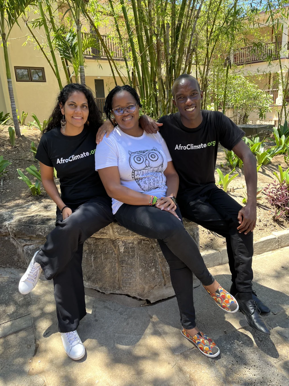 Nassima Sadar-Gravier, a woman with a medium skin tone sits to the left of Susan Mbalu, a Black woman with a deep skin tone, and Joseph Nguthiru, a Black man with a deep skin tone. Nassima and Joseph are wearing shirts that read, “AfroClimate.org.”