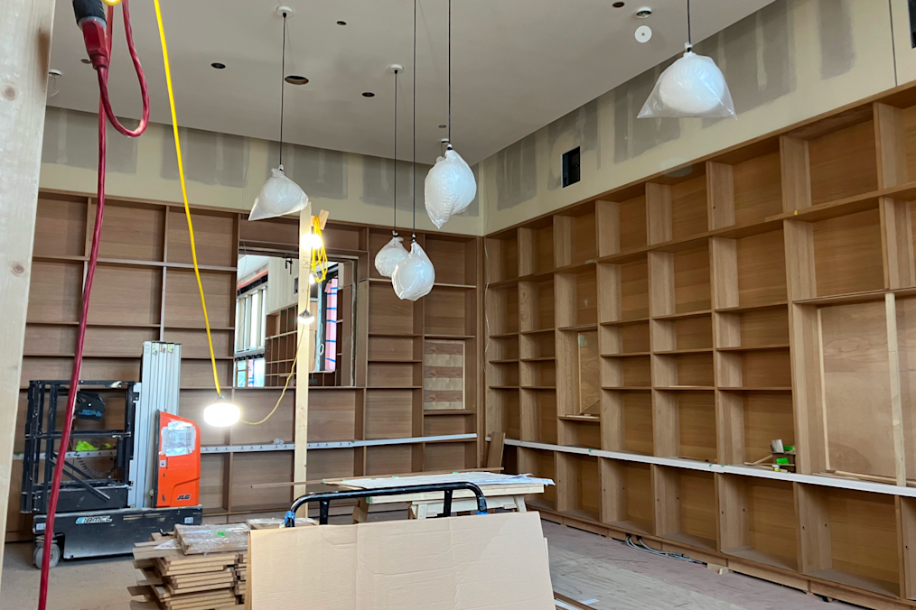 An image of the President's Reading Room in the Chicago Public Library located on the Obama Presidential Center campus. The picture features several brown bookshelves leaning up to the ceiling. On the ceiling are several white pendant lights, hanging. On the floor is a small black crane and several wood beams stacked on the floor.