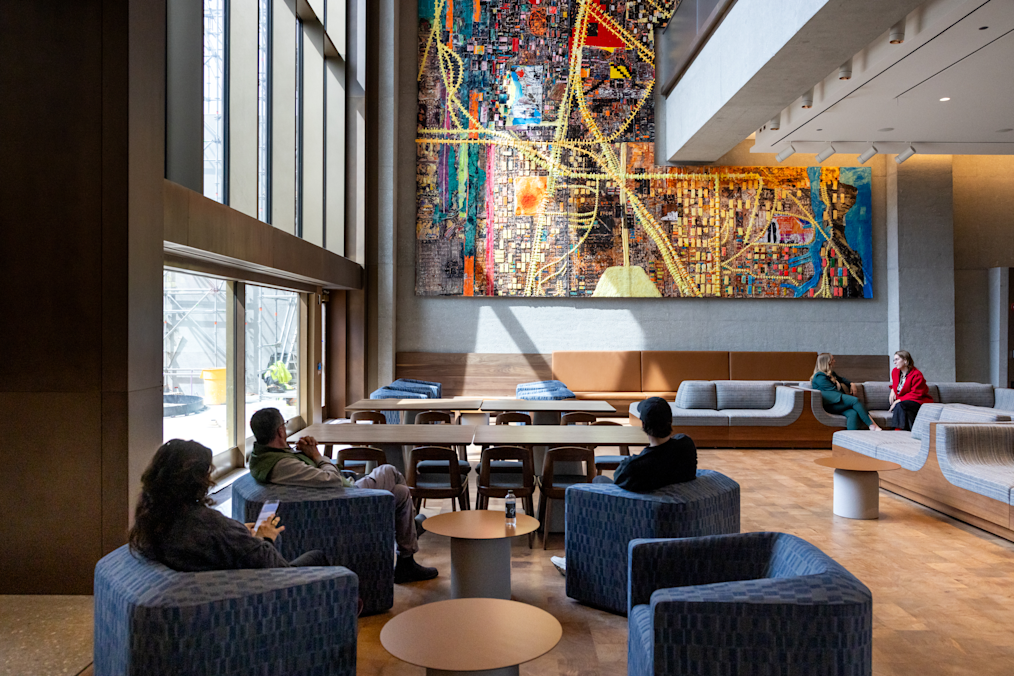 A high-angle photo shows a spacious, sunlit Our Story Atrium with light wood floors and grey concrete walls. Floor-to-ceiling glass windows line the left side, letting in natural light that casts soft, angled shadows across the room. On the back wall hangs a large, vertical, multi-colored tapestry featuring grid-like, abstract patterns in shades of yellow, red, blue, green, and black, reminiscent of a colorful city map. The tapestry is called the City of the Big Shoulders, a new installation from Mark Bradford.