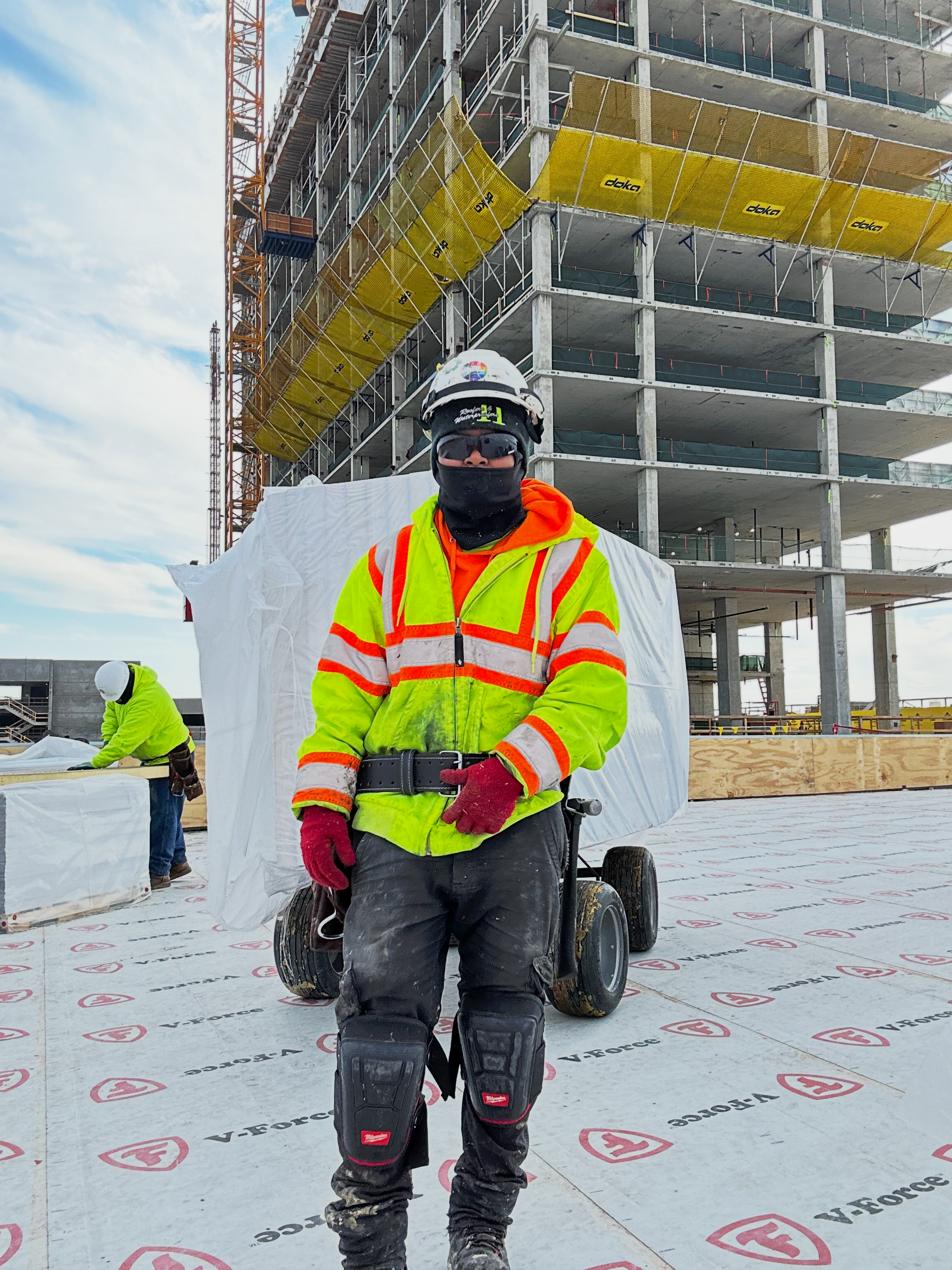 Toryn Parks, a Black woman with a medium skin tone and locs, stands atop of a roof. She is wearing a white hard hat and protective gear. A building structure is in the background.