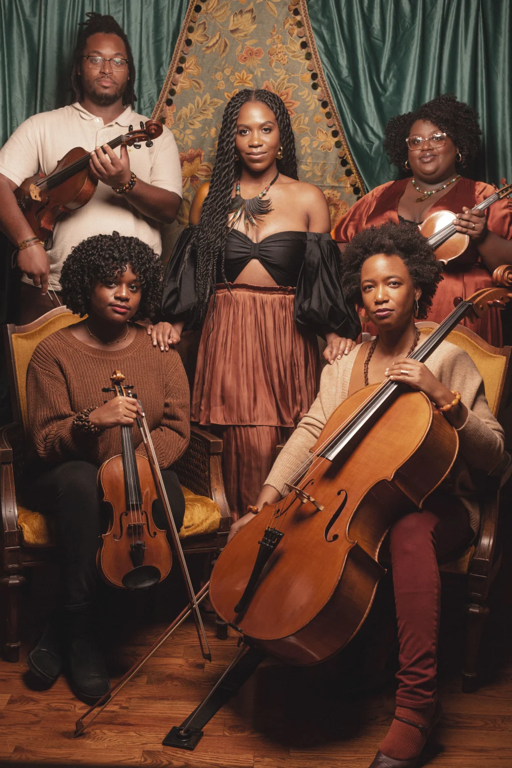 Kori Coleman, a Black woman with a deep skin tone, stands in the middle of a four person string quartet. They are a range of medium to deep skin tones and are holding a viola, violin, and cello. All are wearing shades of brown. 