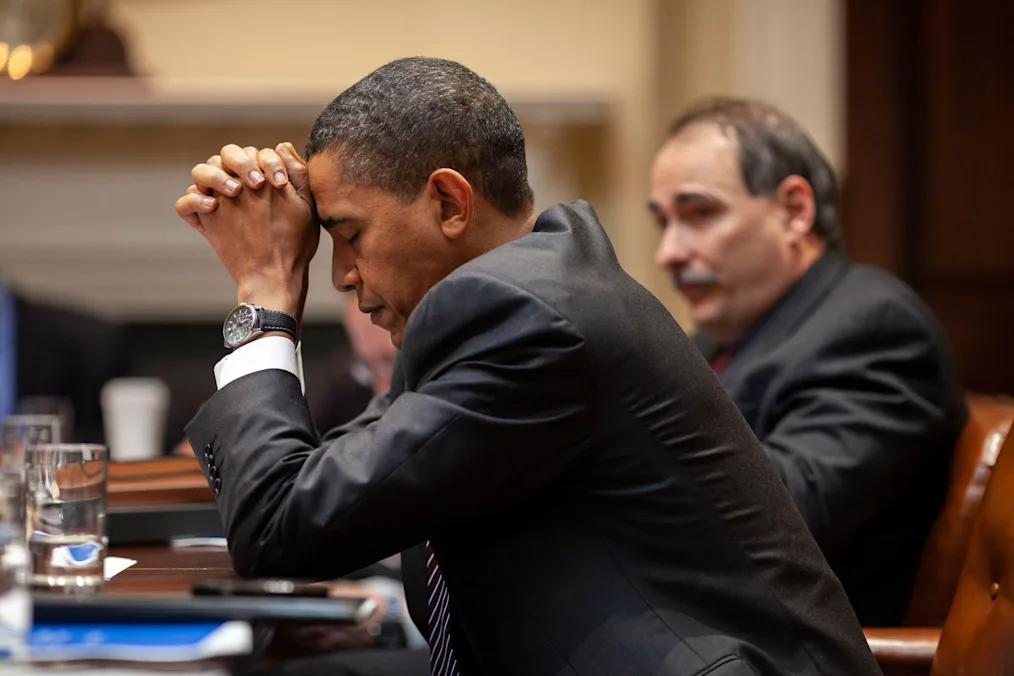 The camera focuses President Obama sits down at a table with his hands together and his head resting upon them while his eyes are closed
