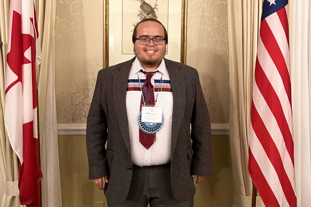 Nick Kennedy, a college-age Native American young man, stands in between a Canadian and American flag on flag poles. He is wearing a brown suit and a white name tag around his neck. 