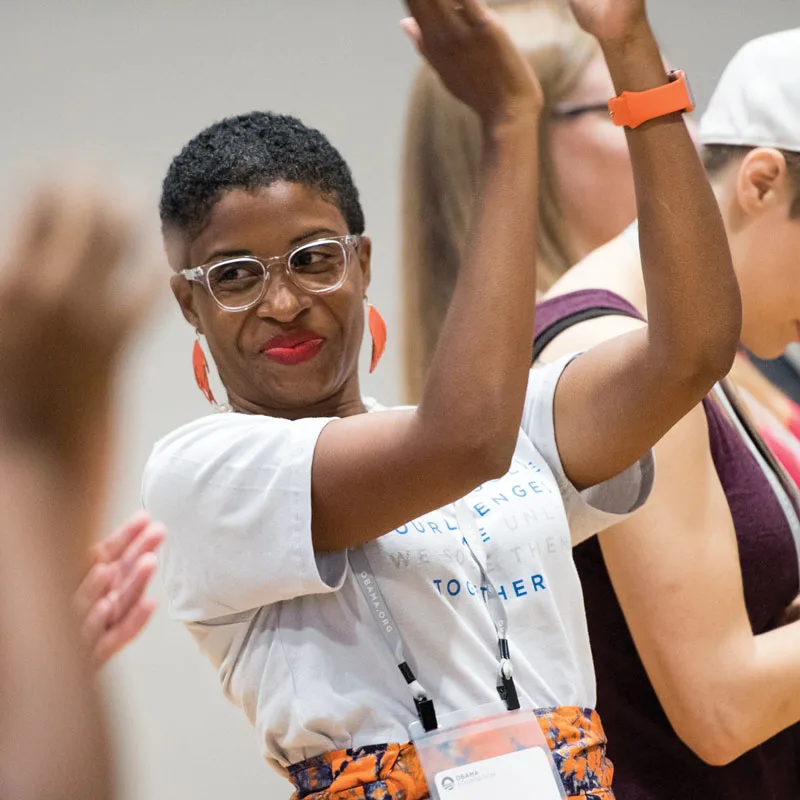A young woman wearing glasses claps over her head during an Obama Foundation Community Leadership Corps kickoff event in 2018.