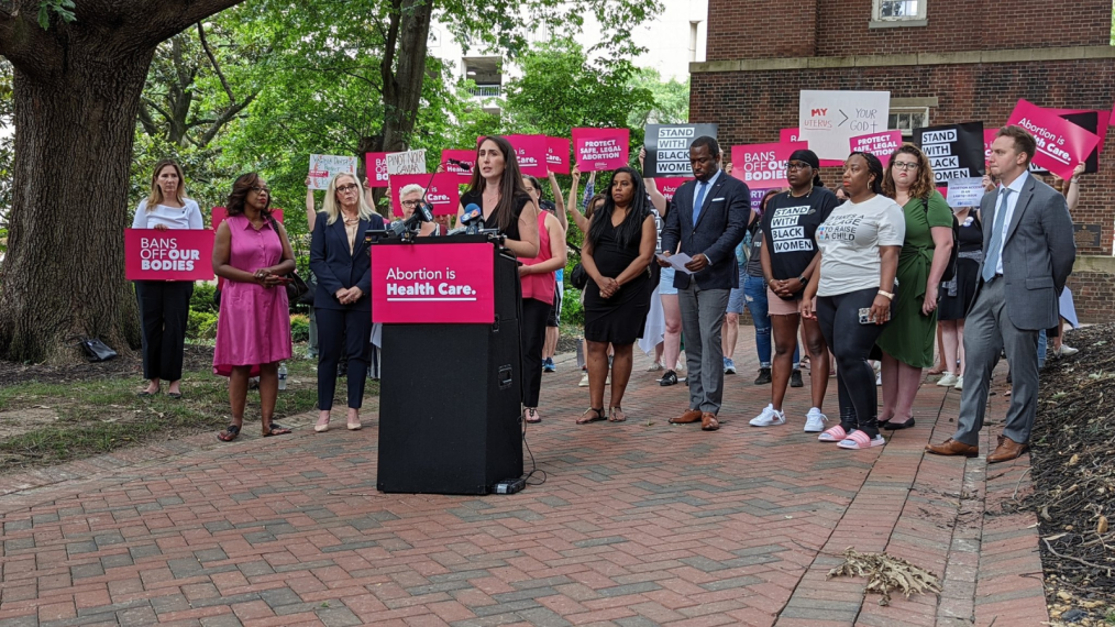 The image is a candid outdoor photo of a press conference the day the U.S. Supreme Court overturned Roe v. Wade with the Dobbs decision. In the photo, dozens of people are standing outside a Bell Tower on a red brick pathway. The people in the picture are holding up Planned Parenthood support signs. In the center of the picture is Jamie Lockhart. Jamie is tall with pale skin and long dark brown hair. She is wearing a black dress. Jamie is standing at a black podium talking into a microphone. Hanging on the front of the podium is a pink rectangular sign that says, “Abortion is Health Care.” 