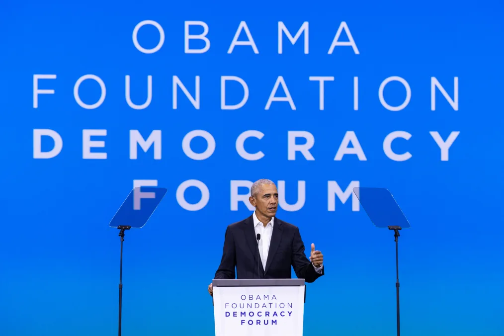 President Obama stands behind a podium and speaks to an audience. A screen behind him reads, “Obama Foundation Democracy Forum.” He is wearing a black suit without a tie.