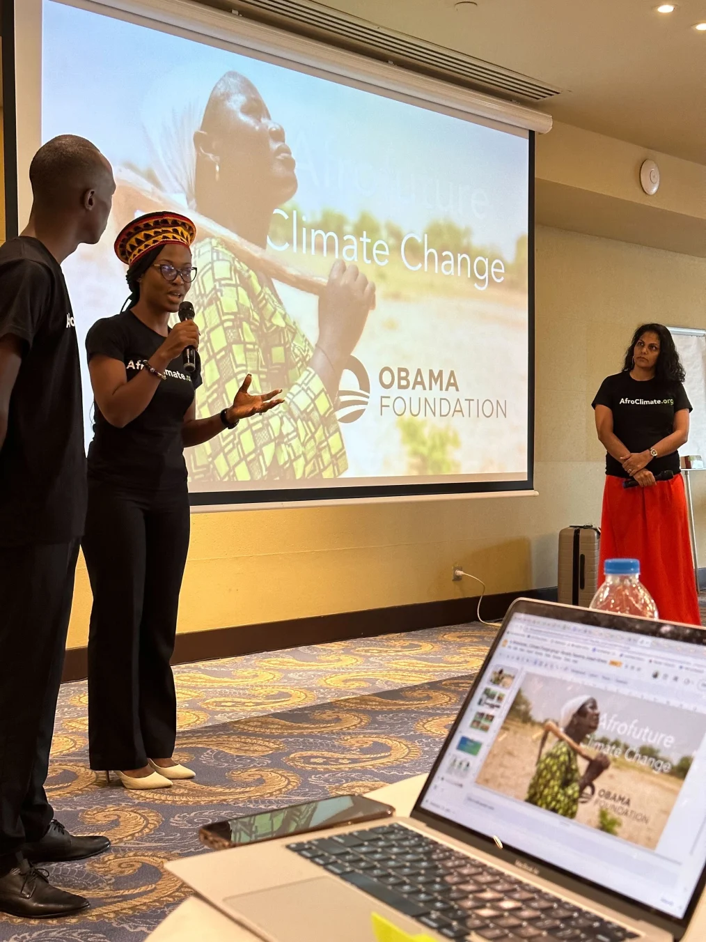 Beverly Ndifoin Niyang, a Black woman with a deep skin tone holds a microphone as she presents in front of a screen that reads, “Afrofuture,” “Climate Change,'' and “Obama Foundation.” On her right is Joseph Nguthiru, a Black man with a deep skin tone. To her left is Nassima Sadar-Gravier, a woman with a medium skin tone. All are wearing shirts that read, “AfroClimate.org.”