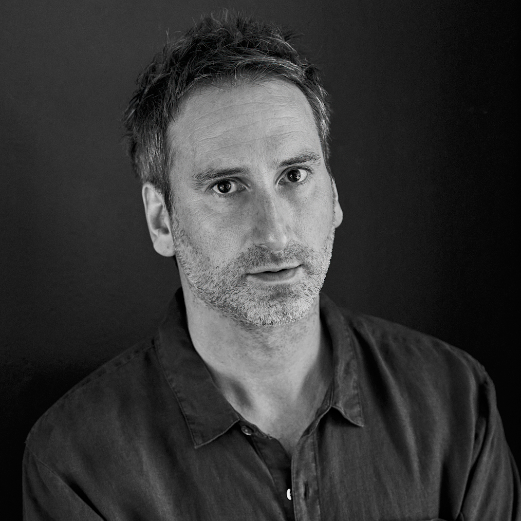 A photographed portrait of Idris Khan. The image is in grey scale. The background of the image is a dark wall. Idris is standing with his arms folder across his chest, staring into the camera. He has a light complexion, short dark hair, and a light colored five-o-clock shade on his face. He is wearing a dark colored button-down shirt with a color. The shirt has a pocket on the left side and white buttons.