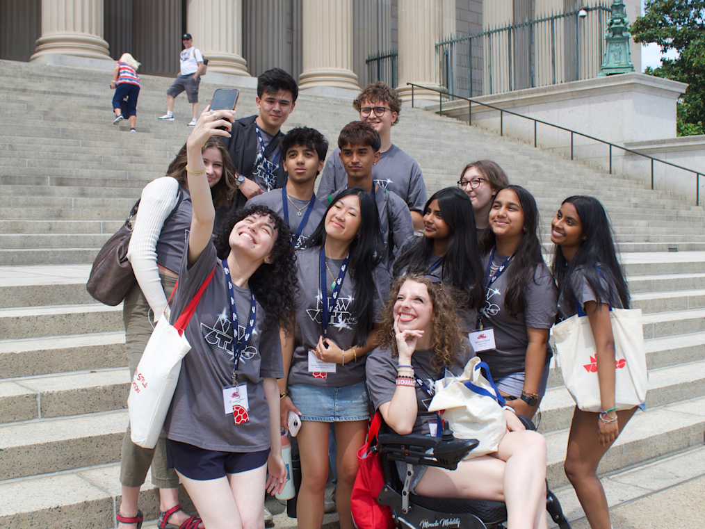 A high-angle photo shows twelve young people with light, medium-light, medium, and medium-dark skin tones smiling on the wide stone steps of a classical building. In the foreground, one person takes a high-angle selfie with a phone, while another sits smiling in a motorized wheelchair. Most wear matching gray t-shirts