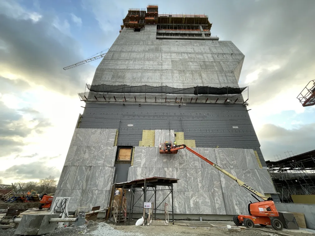A low angle shot shows workers on an orange basket lift affixing a panel of “Tapestry” stone to the lower west face of the Museum Building. A cloudy sunny sky is in the background. 