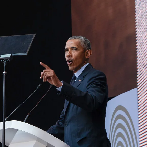 Barack Obama stands at a podium, mouth open and hand raised in a pointing motion. A teleprompter can be seen in front of him.
