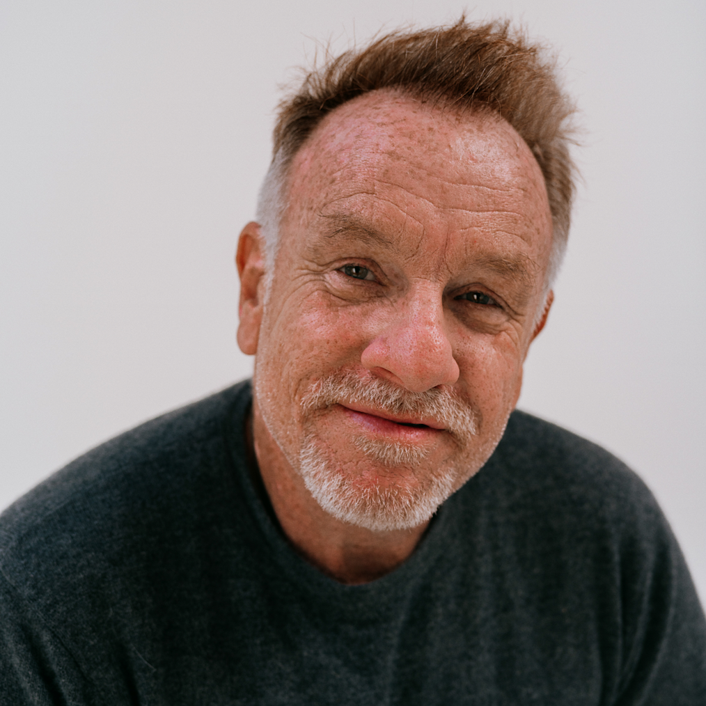 A photographed portrait of Jack Pierson. Behind Jack is a white wall. Jack is staring into the camera. He has an olive complexion, short light colored hair, and a goatee. He is wearing a dark colored sweater.