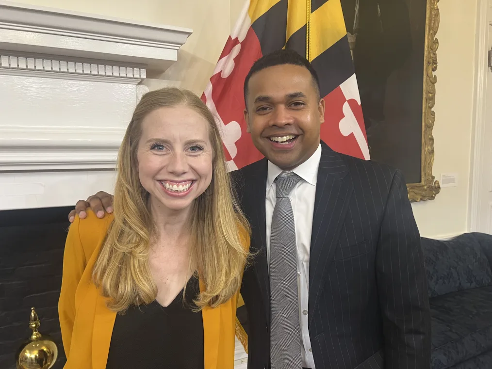 44 Alumni, Tess Hetzel (on the left) and Paul Montiero Jr. (on the right) are smiling into the camera. They are located in an office in the Maryland Capitol. In the room is a white fireplace with a black firepit, a black lace couch, and a beige wall with a larger painting – person unseen – with a gold frame. Tess Hetzel has a pale complexion, long reddish blonde hair, and blue eyes. She is wearing a yellow cardigan and a black v-neck t-shirt. Paul Montiero Jr. has a brown complexion, dark brown curly hair, and brown eyes. He is wearing a black and white pinstriped suit, with a white button-down, and a gray tie. 