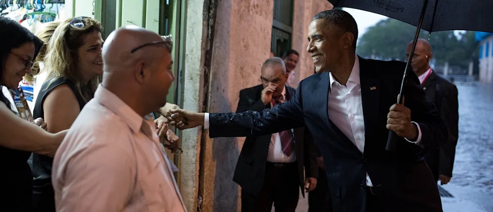 President Obama smiles holding an umbrella in the rain shakes hand with a woman with a light skin tone surrounded by a group of people. 
