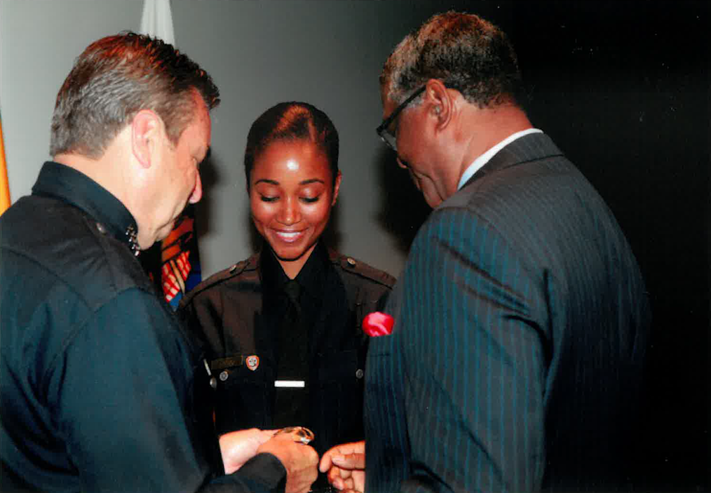 Capt. Shannon White, a Black woman with a medium skin tone, smiles as she’s pinned by her father Capt. Paul Enox and Jim McDonnell, chief of police for the Los Angeles Police Department.