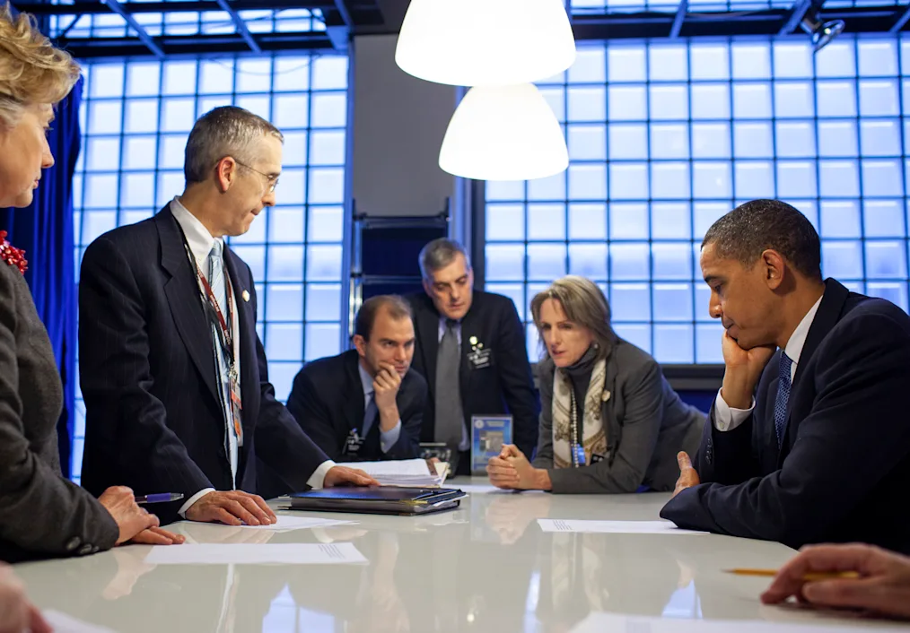 President Obama sits at a long white counter looking over a document with other older men and women with light skin tones. They are sitting around him in business attire 