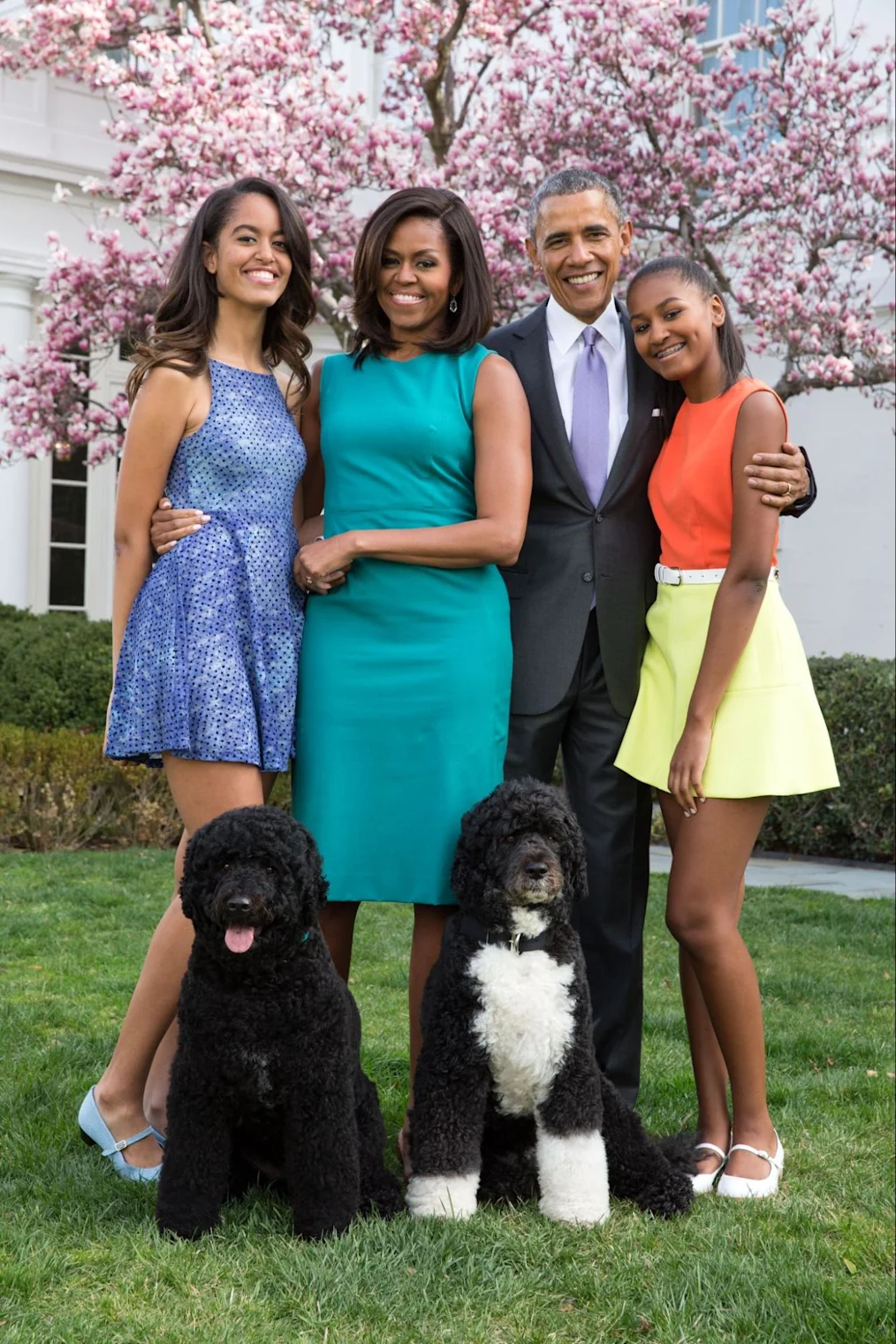 The Obama Family stands and poses for a picture in the Rose Garden of the White House. From left to right is Malia Obama, Michelle Obama, Barack Obama, and Sasha Obama. Sitting in front of the family are Bo and Sunny.