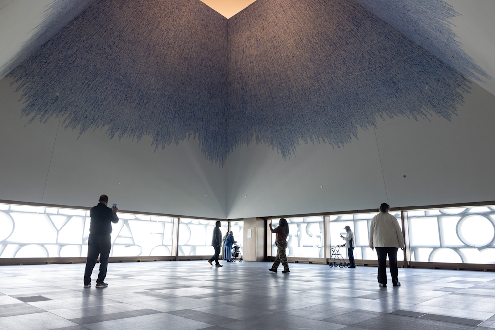 A wide horizontal photograph captures a brightly lit interior of the Sky Room located in the Obama Presidential Center Museum Building. The Sky Room has a tall, inverted-pyramid-style ceiling covered in a mural called Sky of Hope by Idris Khan. The mural is a dense, cascading pattern of blue lines and hand-stamped text that grows thicker toward the square peak at the very top, resembling an inverted whirlpool of blue ink. Along the lower perimeter of the room, wide, low-set horizontal windows frame cut-out letters that form words against a bright outdoor backdrop. Seven people are spread out across the polished gray-tiled floor.