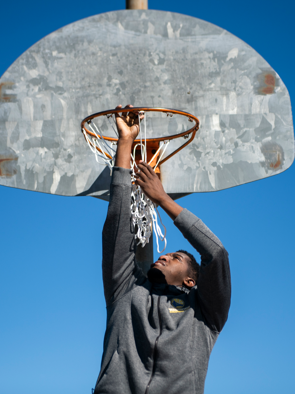 A Black man wearing a sweatshirt holds onto the rim of a basketball net, looking upward. The sky behind is a deep blue.