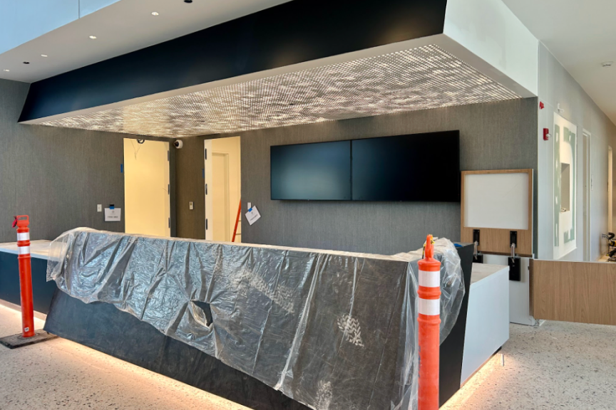 A photograph of the Welcome Desk located in the Lobby of Home Court at the Obama Presidential Center. The desk is covered in plastic and there are two orange cones on either side. Above the desk is a rectangular light fixture with several small lights. On the wall behind the desk are two blank digital screens.