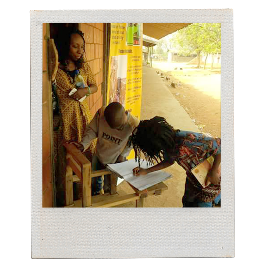 A Polaroid-style photo of two Black children looking over a notebook while one holding a book writes in it. A Black woman stands in a doorway looking at them. Behind them is a brick building, vertical banner and concrete pathway surrounded by dirt.