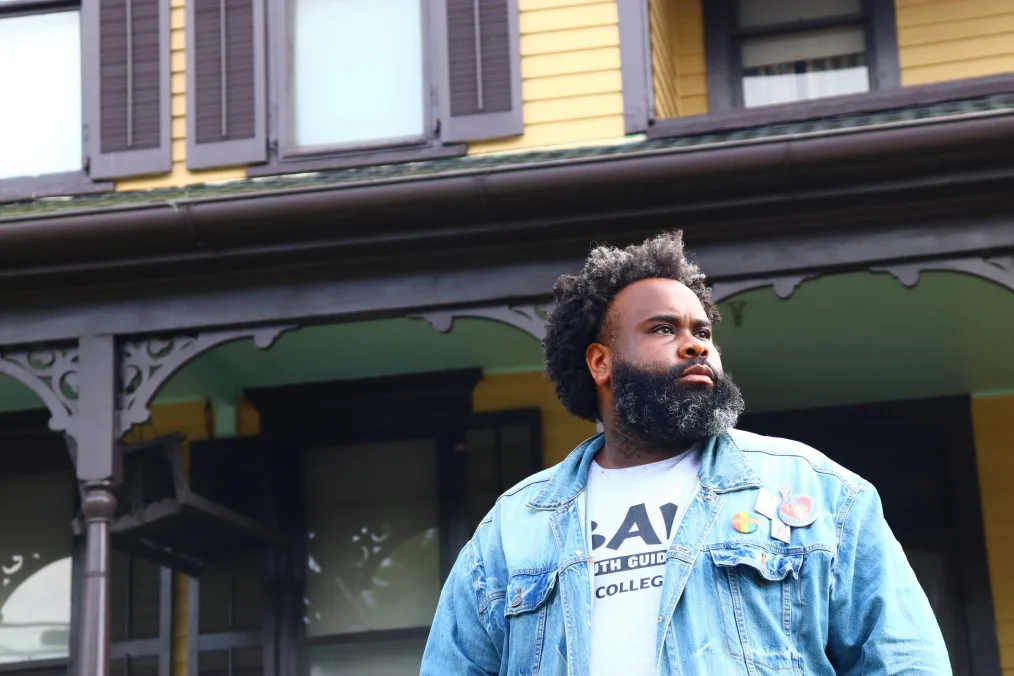 DeKevious Wilson, a Black man with a deep skin tone, looks off camera as he stands in front of a home. He has a salt and pepper afro and beard. He is wearing a Becoming a Man t-shirt and denim jacket. 