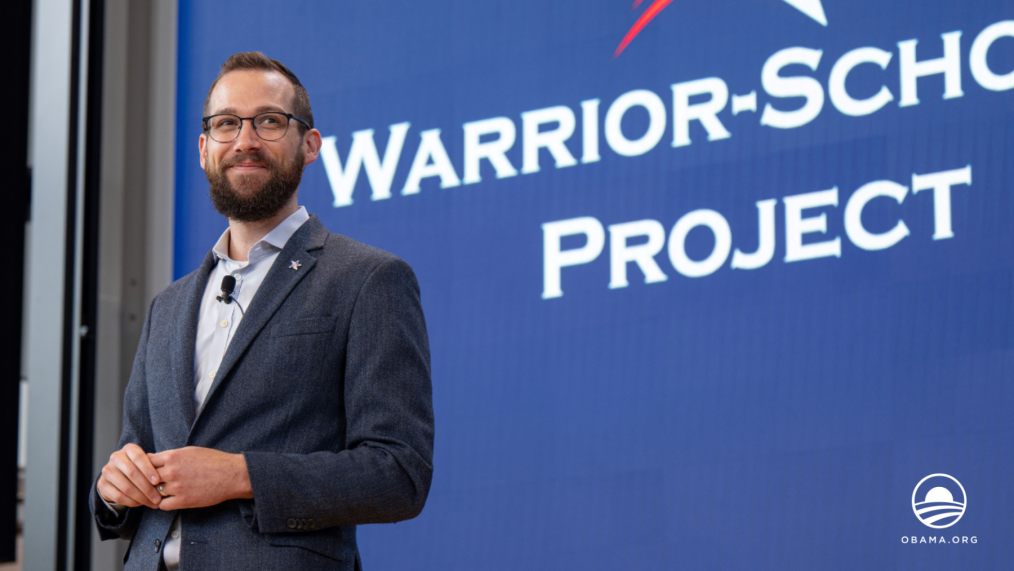 Ryan Pavel, a man with light skin tone, stands smiling in front of a sign that reads "Warrior-Scholar Project."