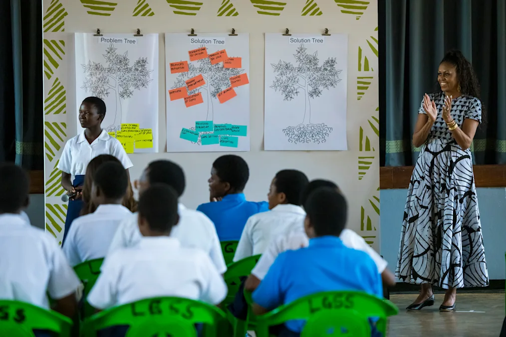 Michelle Obama, a Black woman with a medium-dark skin tone, stands on the right in a pattern-printed dress, smiling and clapping her hands. On the left, a young Black girl in a white shirt and blue skirt holds a microphone in front of three large posters with tree drawings. Other students in white or blue shirts sit in the foreground facing them.
