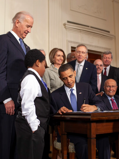 President Obama sits at a desk, pen in hand, as other politicians look on. A young boy at the front, dressed in a suit vest and tie also looks on.
