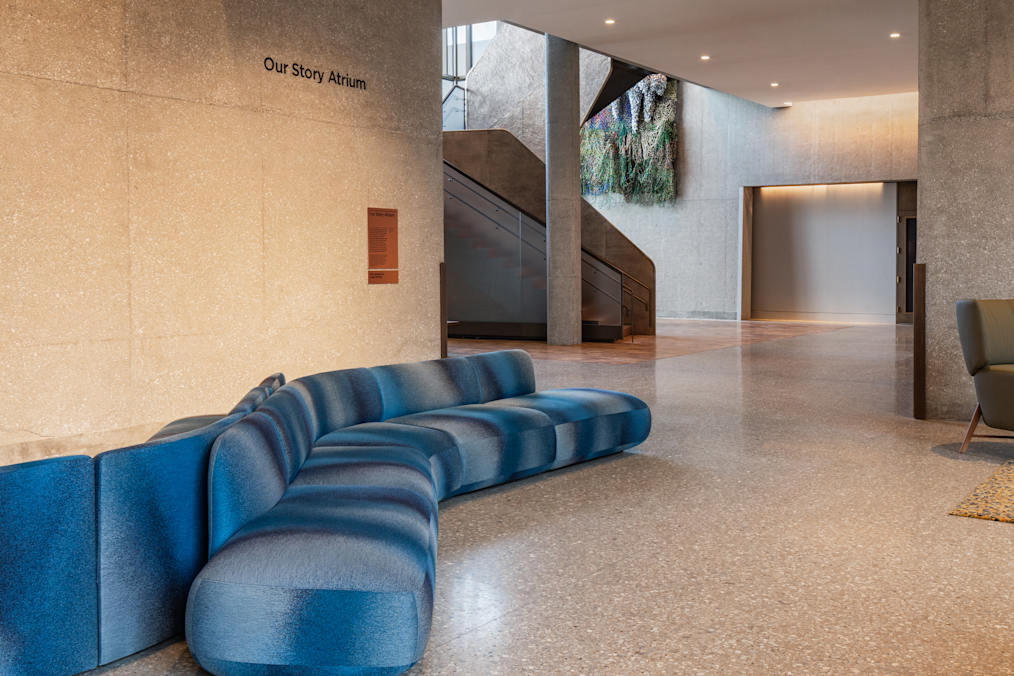 A wide, interior view of a bright, open building space with terrazzo flooring and polished concrete walls. In the left foreground, a curving, low-profile modular sectional sofa upholstered in shaded blue-and-gray fabric sits near a concrete wall. Black lettering mounted high on the wall reads "Our Story Atrium," with a small rectangular sign positioned directly below it.