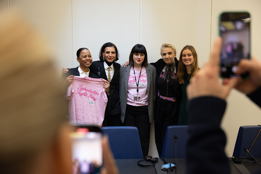 Nika Kovač, a white woman with a light skin tone, poses with a group of people with a range of light to dark skin tones. All are smiling and holding shirts that read “My Voice, My Choice.”