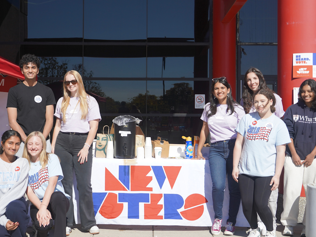 Eight smiling young people stand and crouch around an outdoor information table with a white banner reading "NEW VOTERS". The participants have light, medium-light, and medium skin tones. A drink dispenser, cups, and snacks sit on top of the table in front of large dark glass windows.