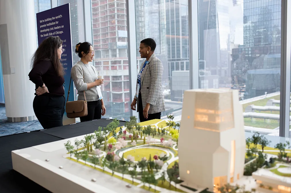 In the background, three women, with a range of light to medium skin tones, are seen conversing. In the foreground there is a small model of the Obama Presidential Center.