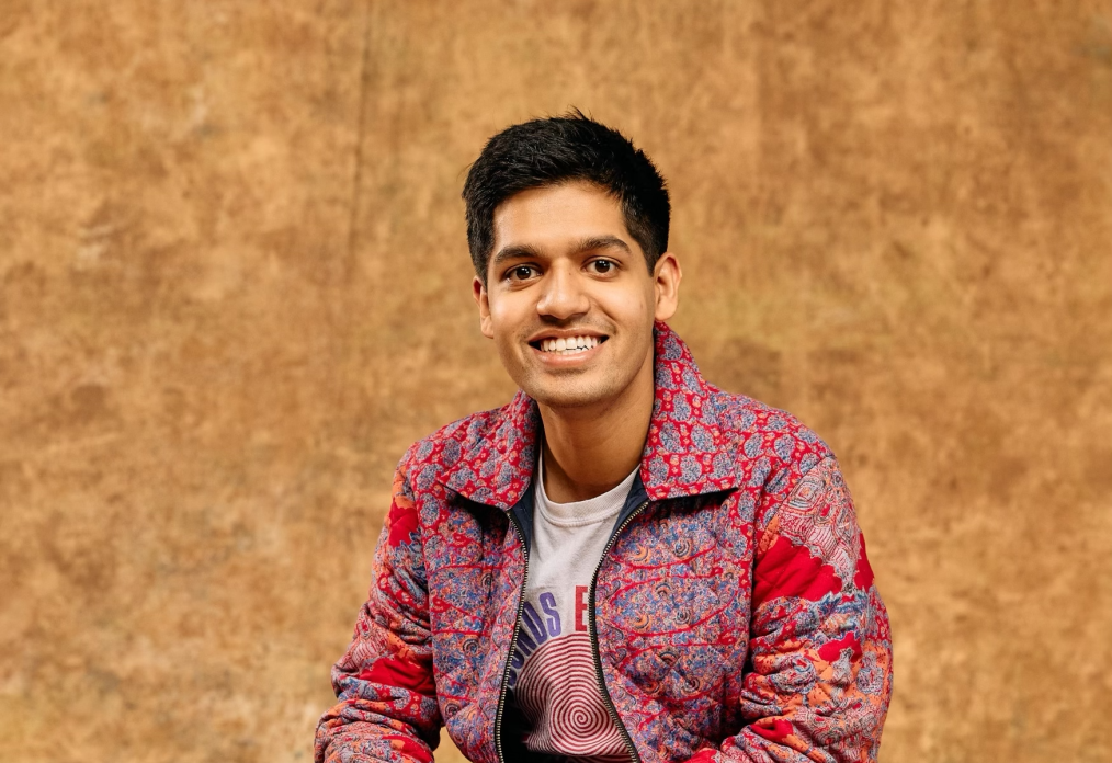 Ziad Ahmed, a man with a medium skin tone, smiles at the camera. He has black hair and is wearing a red patterned jacket. He is sitting on an apple box. The background is brown. 