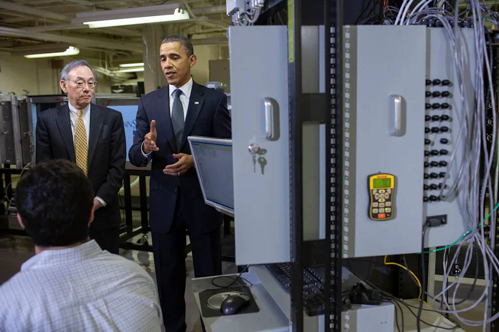 Barack Obama a black man wearing a deep dark blue suit standing next to a man with a light neutral tone wearing a black stripe suit and yellow tie talking to a man with a light skin tone who is sitting down in front of them next to an electricity box with multiple cords on it 