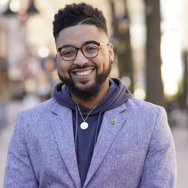 Daniel Fairley II, a Black man with a light skin tone smiles at the camera. He has short black hair and glasses. He is wearing a light purple blazer and a dark purple hoodie.