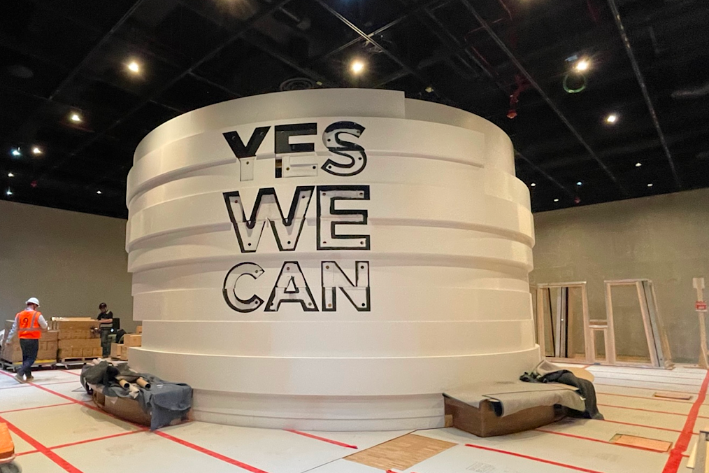 An image of "Yes We Can," an exhibit in the Obama Presidential Center Museum. The exhibit is a circular enclosure that is white. On the exterior of the enclosure are the words "Yes We Can." Above the exhibit is a black ceiling.