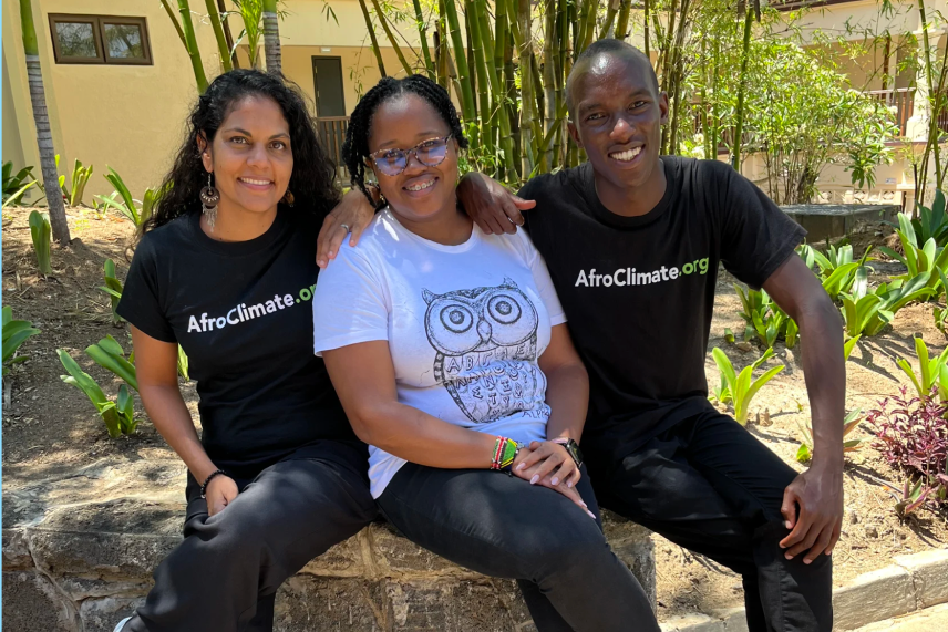 Two women and one man pose for a photo. One woman has medium-dark skin tone, and the other two people have dark skin. Two of them wear shirts with the words "Afroclimate.com." They sit on a rock wall in front of dirt and greenery.