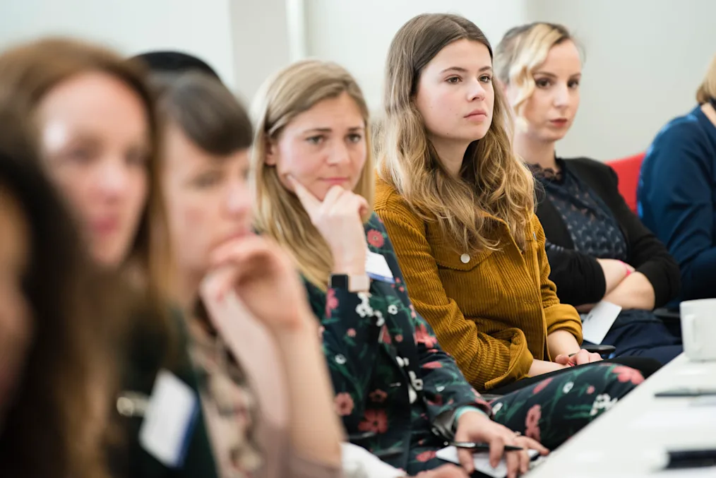 6 women with light neutral tones sitting next to each other at a table making the same serious facial expression but the camera is zoomed in on one woman with brown hair and blonde highlights wearing a mustard corduroy jacket 