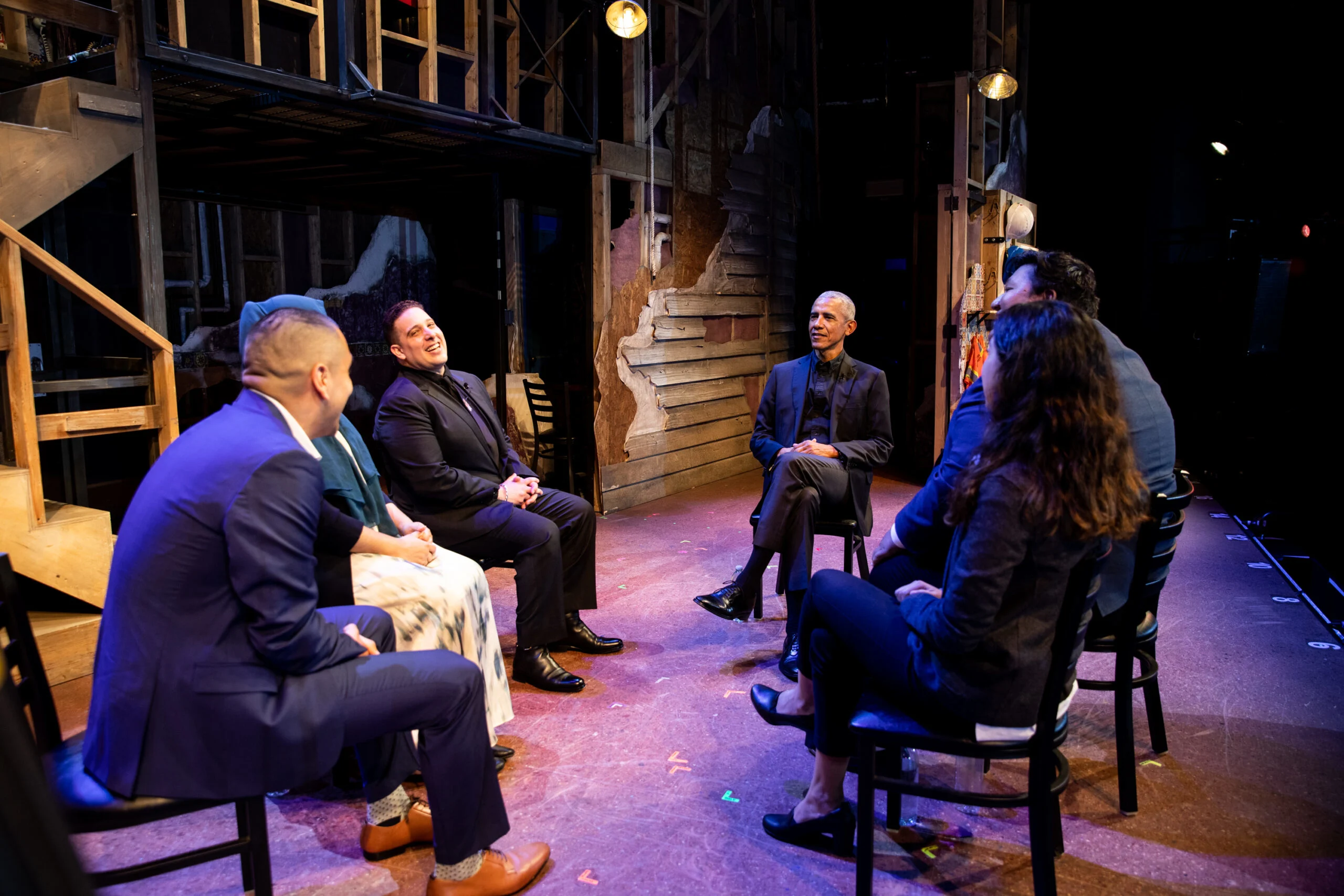 President Obama sitting on stage talking to five other people wearing formal clothing
