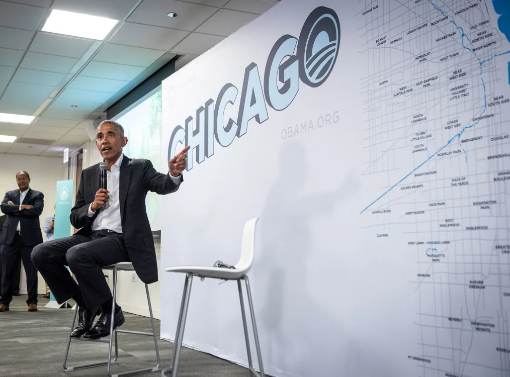 President Obama speaks on a panel. In the background is a board that reads, "CHICAGO." It is a bright blue color with the Chicago map on the right side.