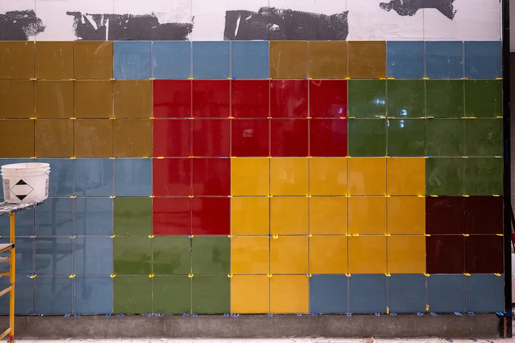 An image of Spencer Finch’s tile artwork, “To Disappear Enhances – (Memory Landscapes).” The artwork is made up of several square tiles on a wall. The tiles are different colors including light blue, yellow, red, green, and different shades of brown.