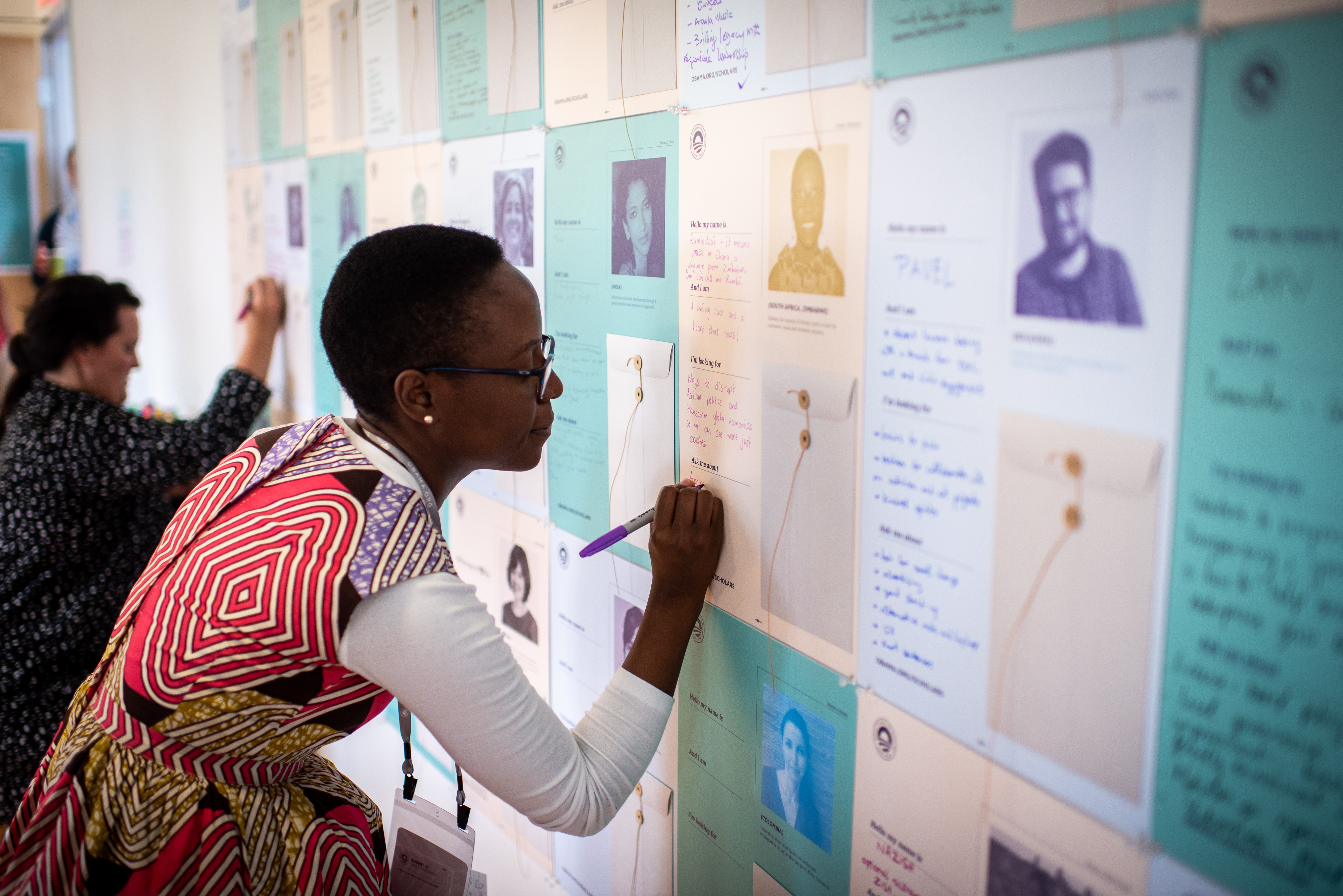 A lady with a deep skin tone wearing a multi-colored dress and a white shirt with 
short black hair and glasses, is writing about herself on a board.