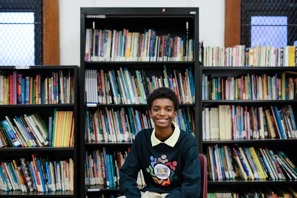 A young boy smiles as he sits in front of a bookshelf. He has a medium skin tone and short afro. He is wearing a yellow polo with a navy crewneck on top. 