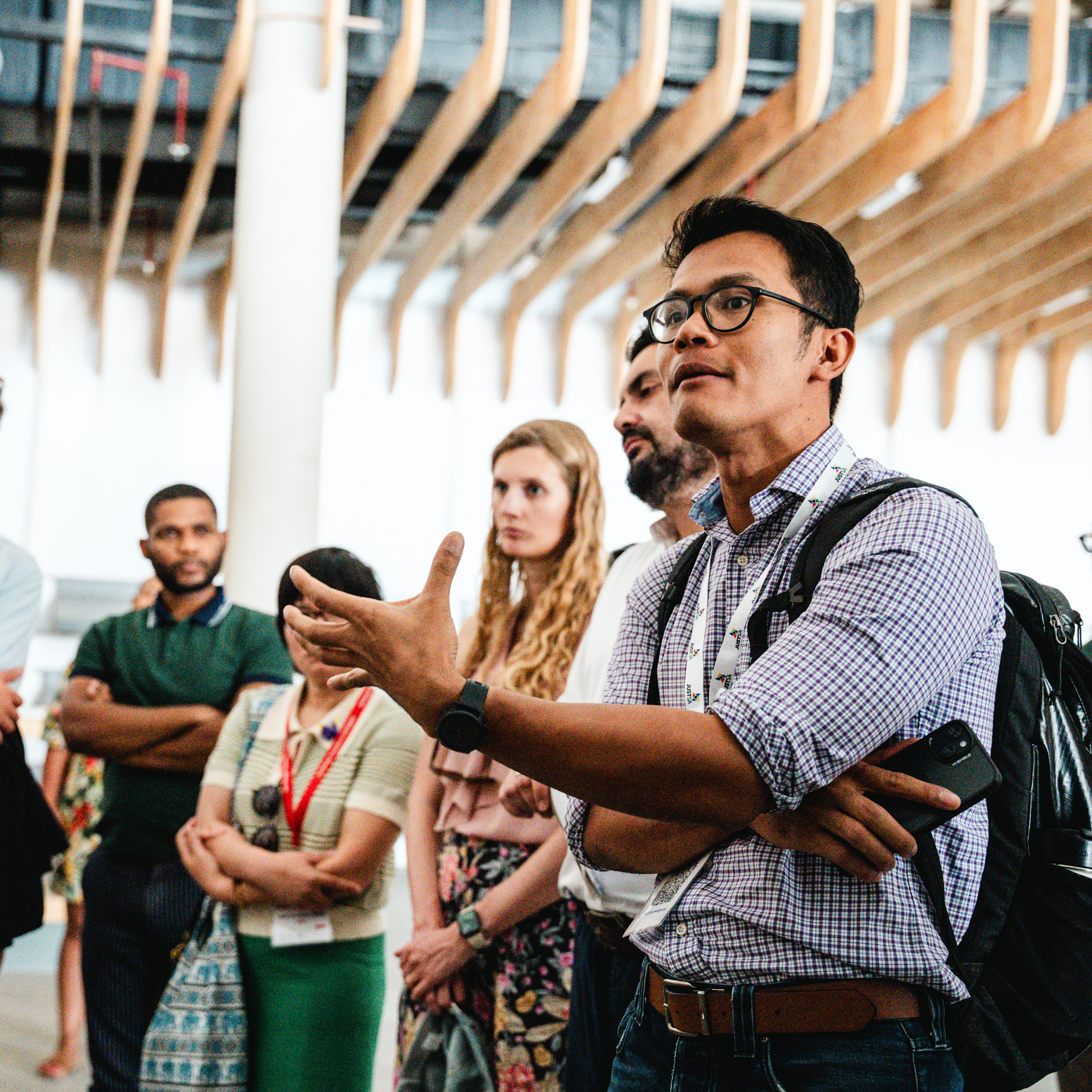 Charles Klaikaew, an Asian man with a light skin tone, speaks in a circle to a group of people with a range of light to medium skin tones. He is wearing glasses and a checkered shirt.