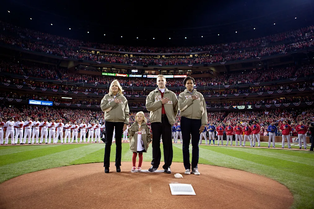 First Lady Michelle Obama wearing a tan jacket and black pants stands on the grass amongst three individuals with light skin tones wearing tan jackets and black pants. In the background there are baseball teams and a stadium full of people with a variety of skin tones. 