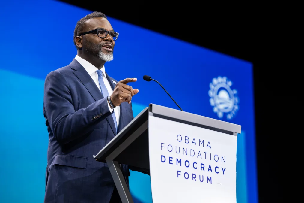 Chicago Mayor, Brandon Johnson, a Black man with a deep skin tone, stands behind a podium as he speaks to an audience. The front of the podium reads, “Obama Foundation Democracy Forum.”