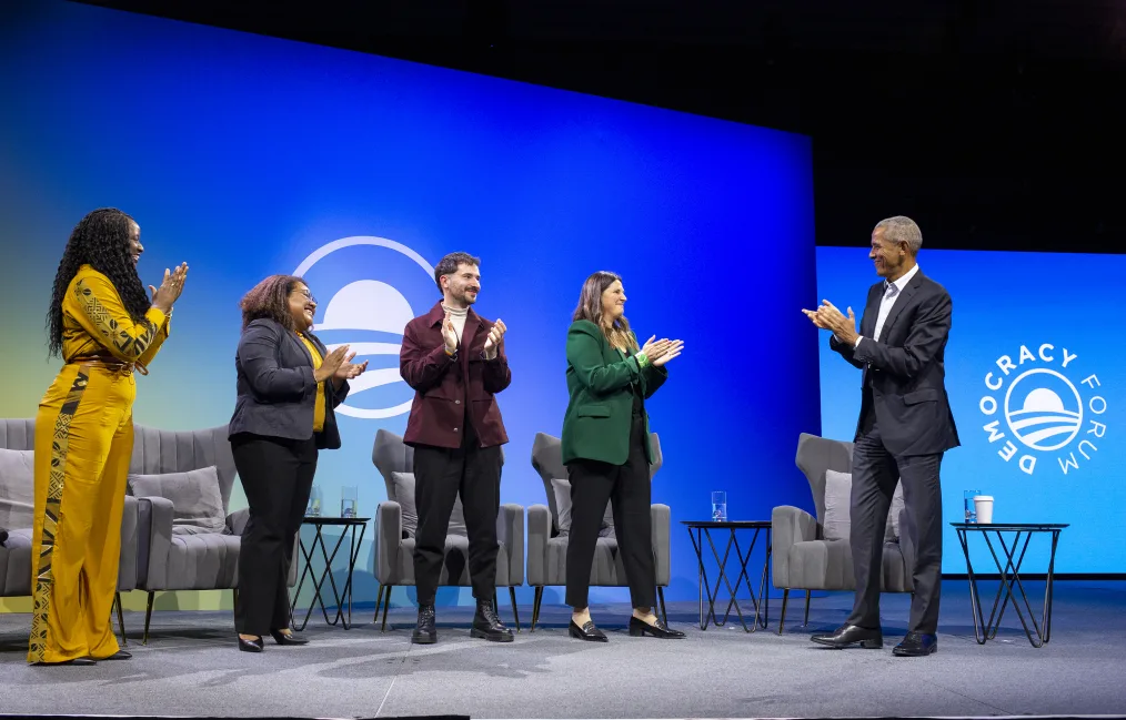 President Obama stands on stage wearing a dark suit and a white button down. He claps towards his right gesturing to four young people with various skin tones wearing business attire. They are also clapping and smiling while gesturing toward President Obama. Behind them, gray wing-backed chairs and a blue and green backdrop with the Obama rising sun logo. 