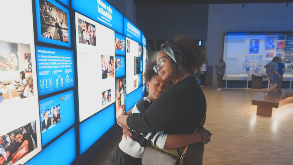 Kendra Robinson, a Black woman with a medium skin tone, and her daughter, a girl with a medium skin tone, hug at one of the Obama Presidential Center Museum exhibits.