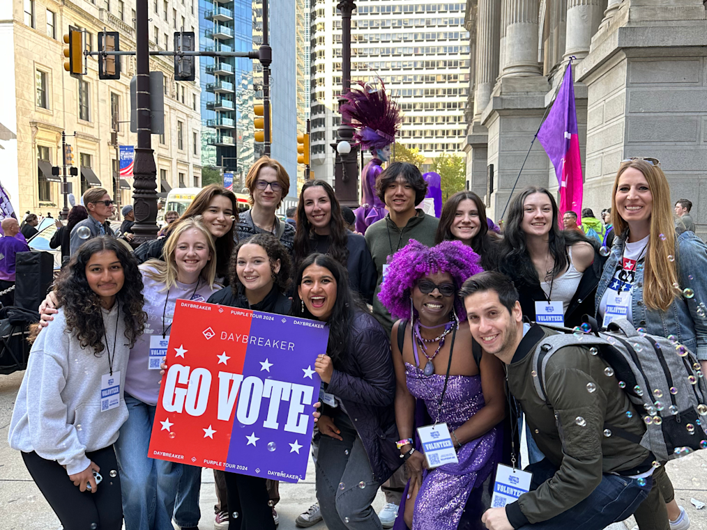 A group of twelve smiling young people stands on a city sidewalk next to a stone building. In the center, two people hold a purple and red sign reading "GO VOTE". The individuals have light, medium-light, medium, and medium-dark skin tones. Soap bubbles float around them under bright daytime light.